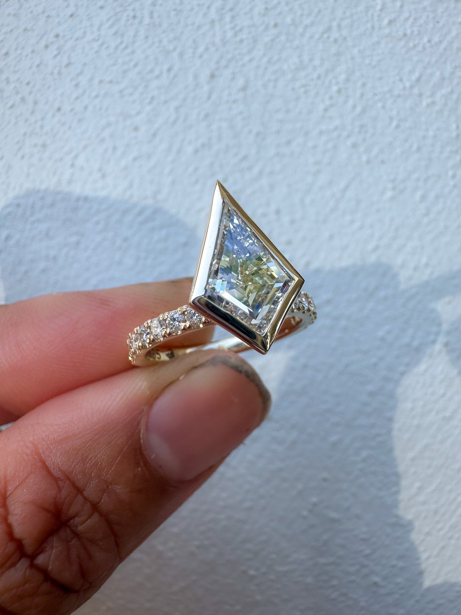 A yellow gold ring with a large kite diamond and a diamond-accented band, held between fingers against a light textured wall.