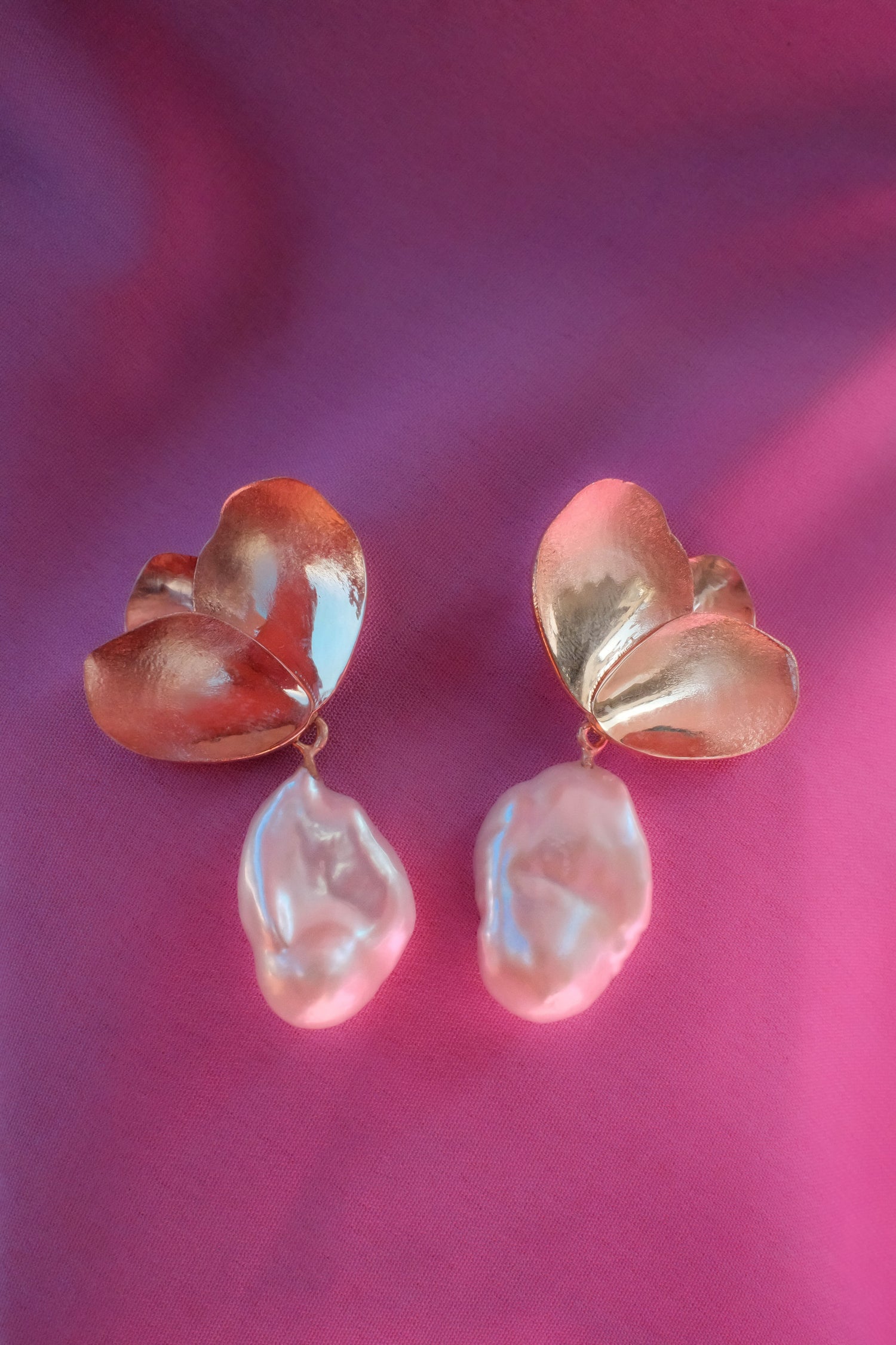 A pair of handmade earrings featuring textured, organic petals made in solid 9K yellow gold with dangling baroque pearls, set against a vibrant magenta background.