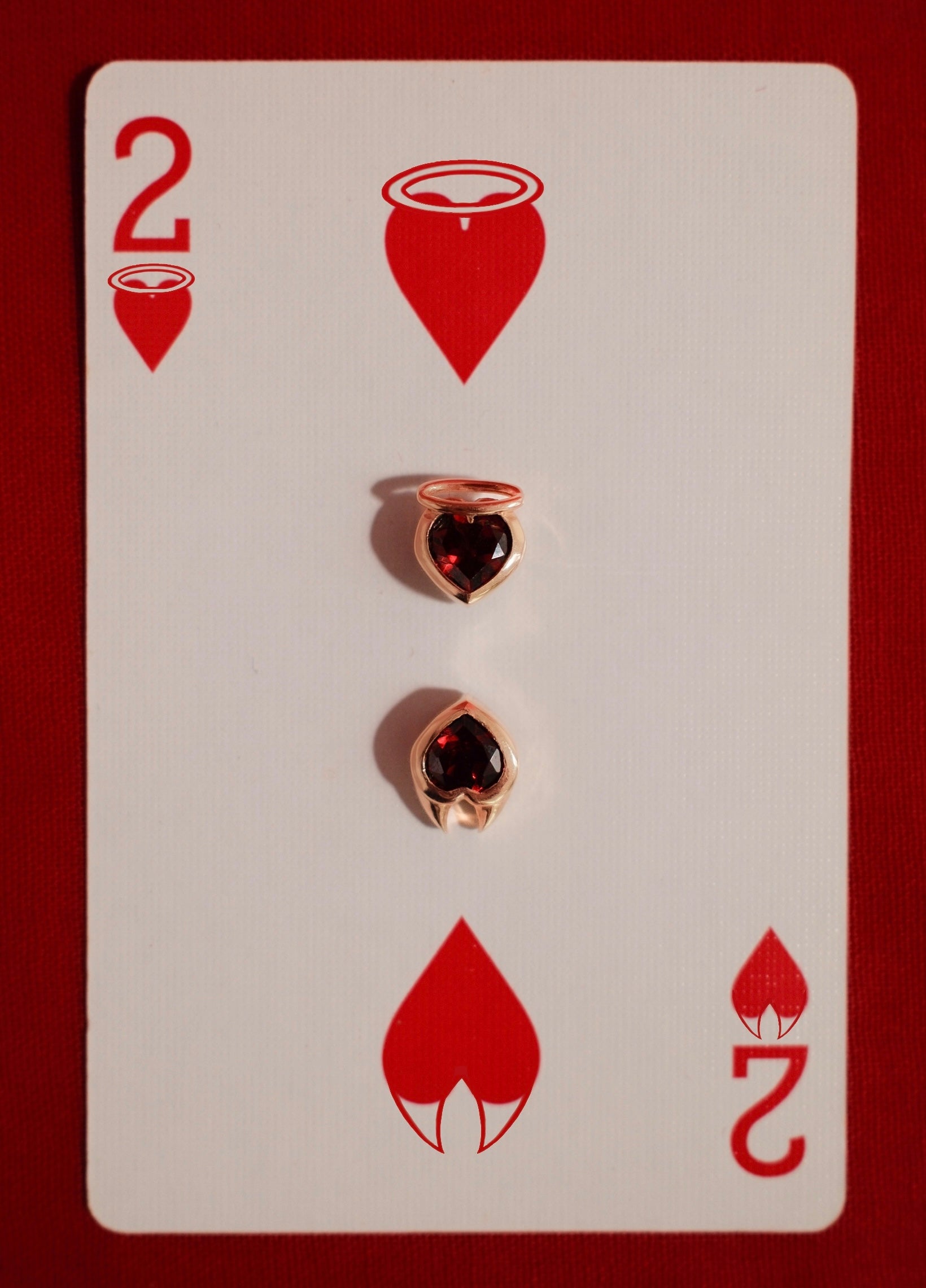 Yellow gold earrings with heart cut garnet gemstones set in an angel and devil style setting on a playing card with red hearts and number 2.