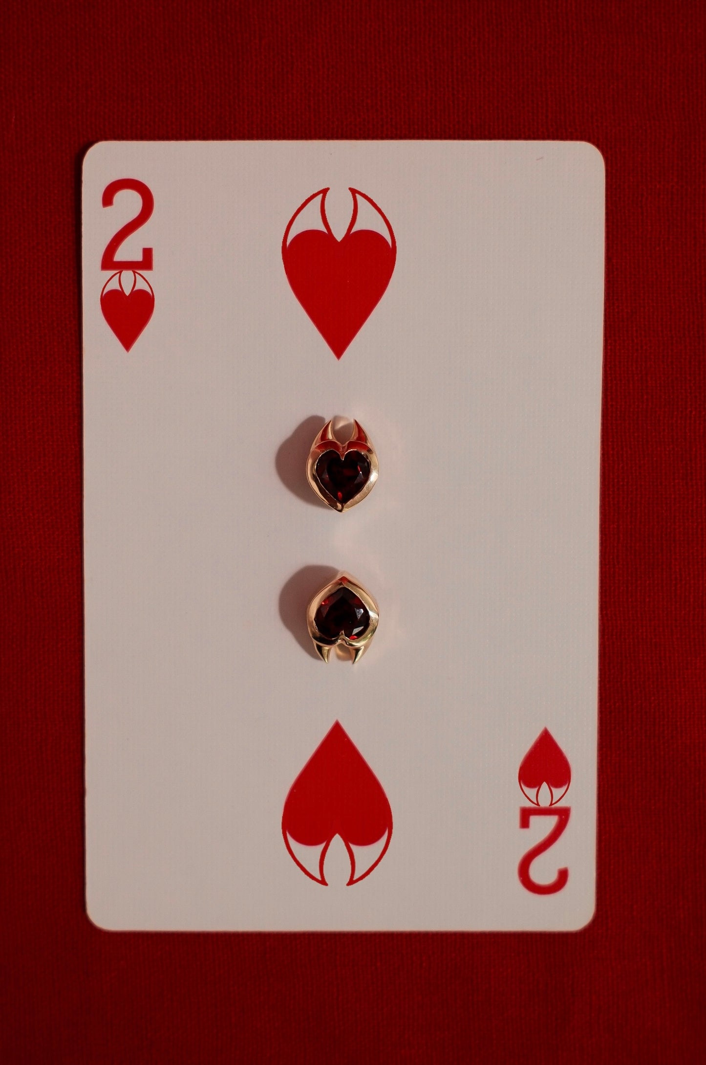 Yellow gold earrings with heart cut garnet gemstones in devil style setting on a playing card with the number 2 and heart symbols.