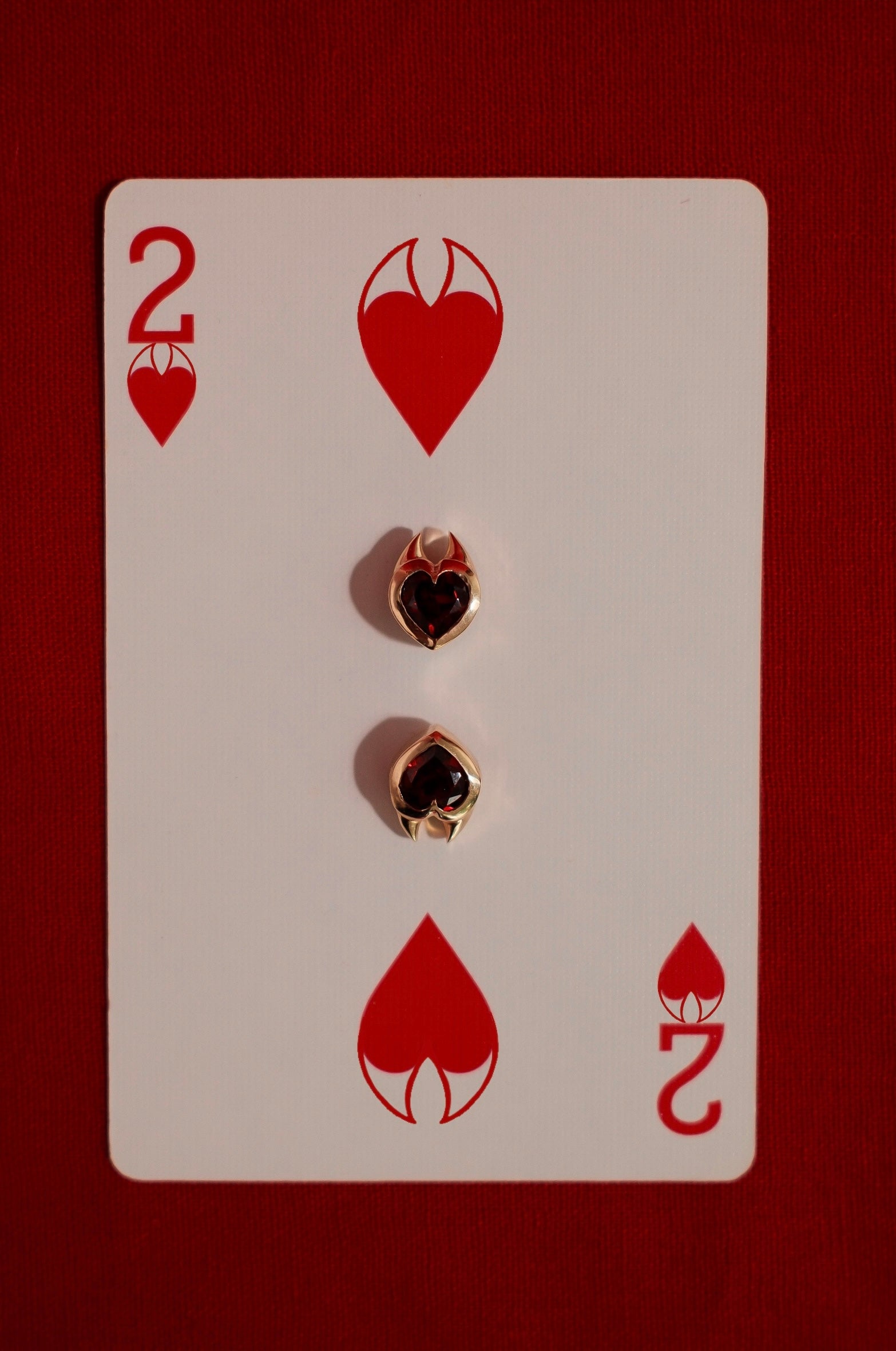Yellow gold earrings with heart cut garnet gemstones in devil style setting on a playing card with the number 2 and heart symbols.